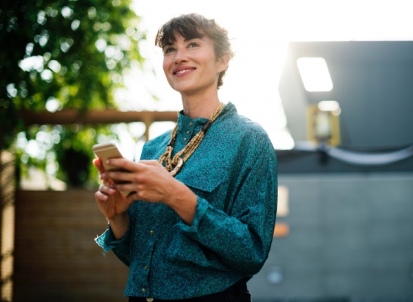 A smiling woman in an azure blouse holding a smartphone while standing outdoors